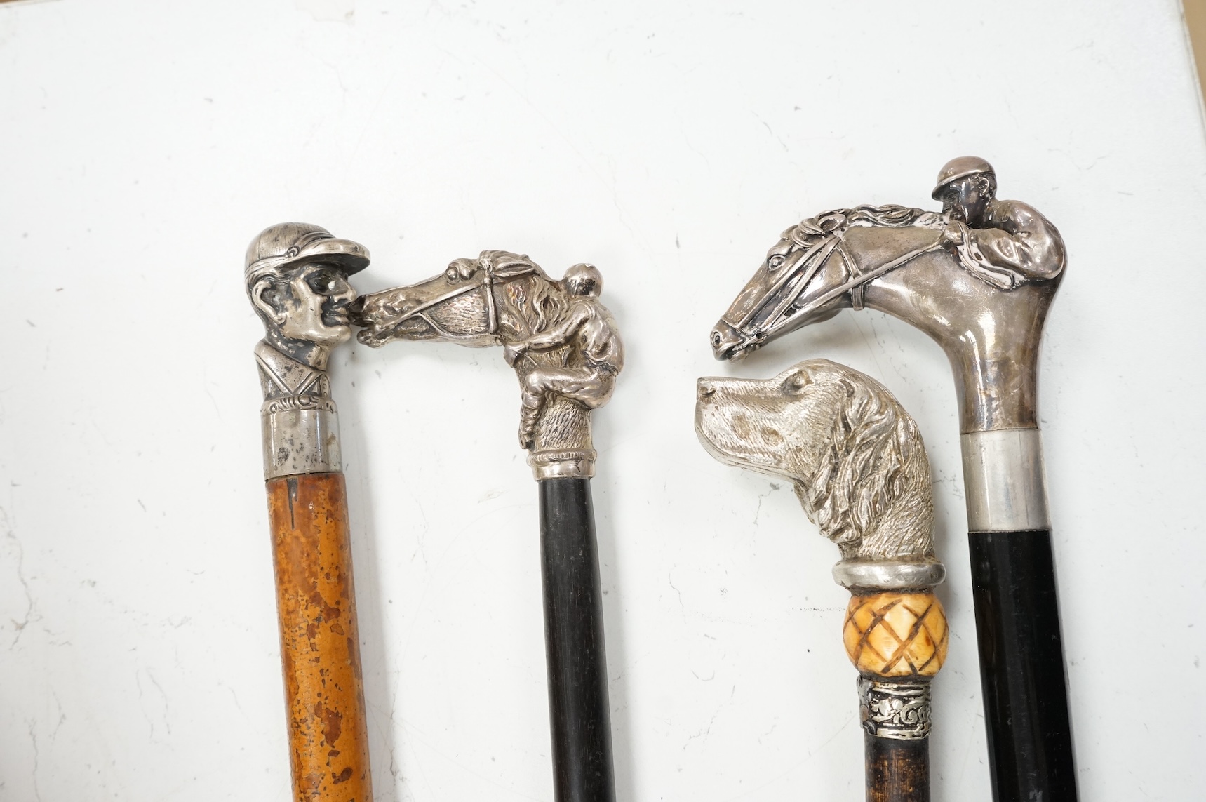 Four 19th century and later walking sticks with silver and white metal tops, to include three horse racing interest examples, largest 96cm in length